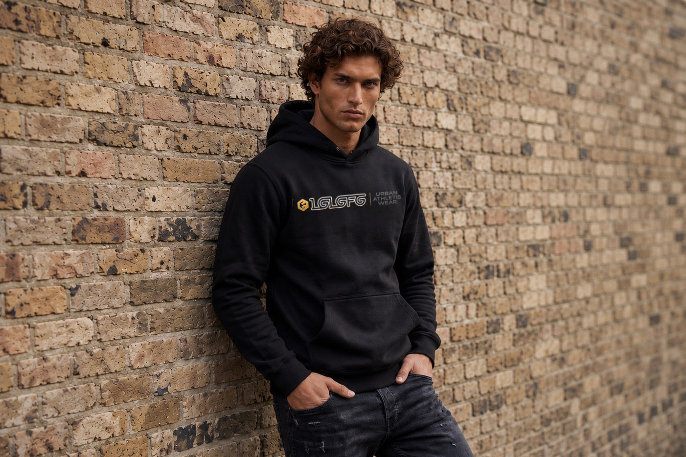 A man wearing a black pullover hoodie with a yellow and white 'OLAFIT' logo on the chest, leaning against a brick wall.