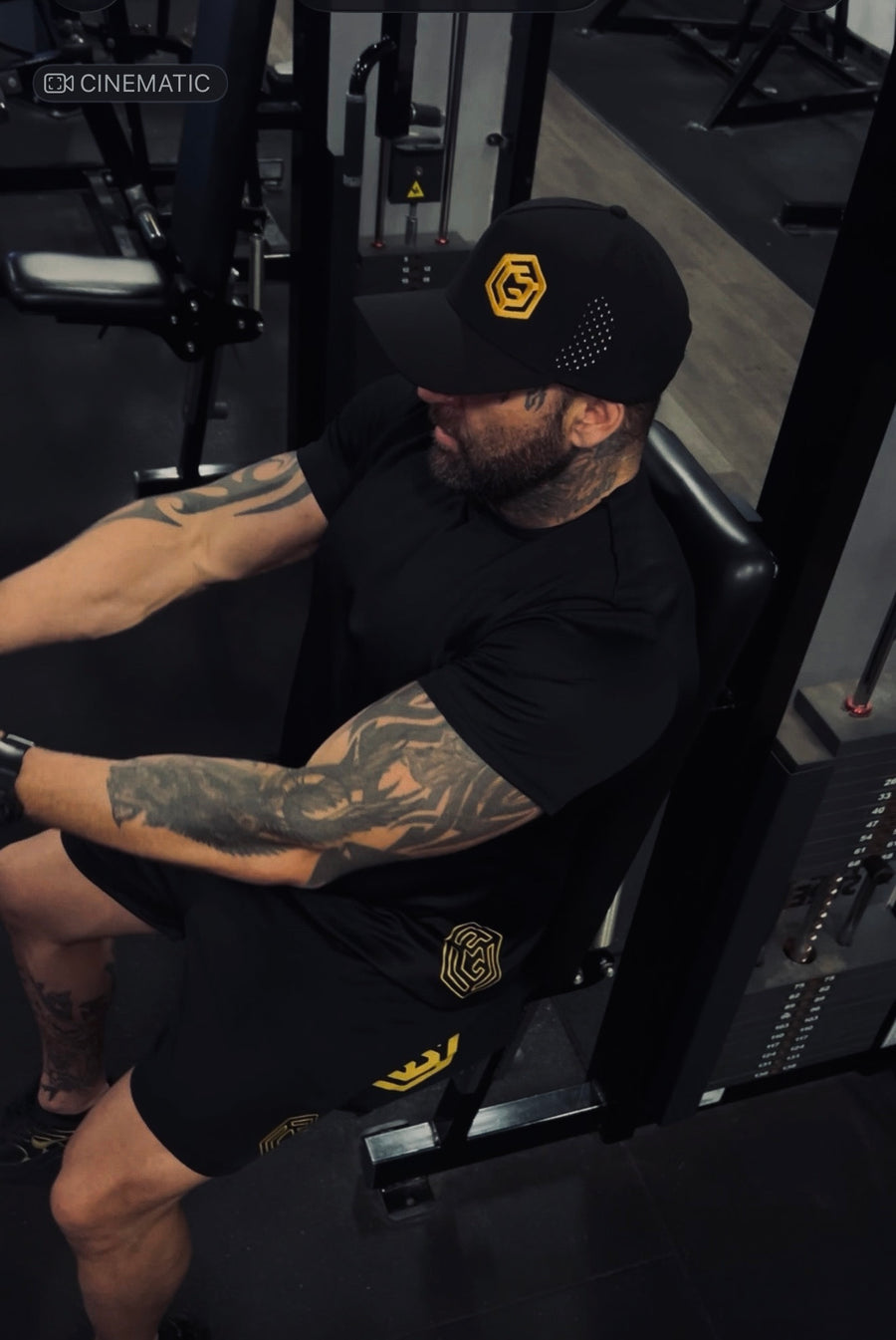 Man wearing a black LGLGFG T-shirt and gold-logo cap on a gym machine